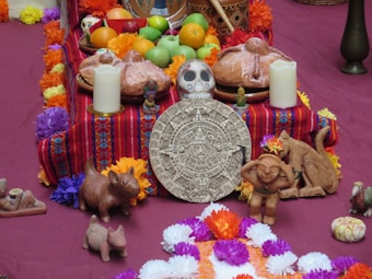 An altar with a variety of traditional items arranged for a cultural celebration. The setup includes fruits such as oranges and apples, decorated breads, colorful marigold flowers, and candles. There are also small figurines and a central focus on an Aztec-style stone calendar. The surrounding vibrant papel picado (cut paper decorations) feature rich colors like red, orange, purple, and yellow.