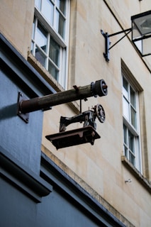 An antique sewing machine is mounted on a wall bracket outside a building with a beige stone exterior and white-framed windows. The sewing machine is weathered and appears to be used as a decorative sign.