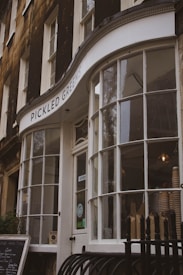 A storefront with large curved glass windows and a sign reading 'PICKLED GREENS'. The building has a classic design with dark exterior walls and white window frames. Inside, there are visible stacked paper cups and a lit hanging bulb, suggesting a cozy, cafe-like atmosphere. A chalkboard menu is positioned outside.
