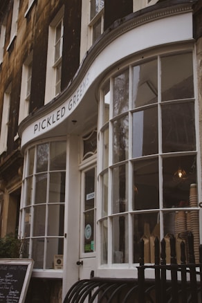A storefront with large curved glass windows and a sign reading 'PICKLED GREENS'. The building has a classic design with dark exterior walls and white window frames. Inside, there are visible stacked paper cups and a lit hanging bulb, suggesting a cozy, cafe-like atmosphere. A chalkboard menu is positioned outside.
