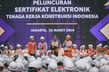 A group of people wearing construction helmets and safety vests gather on stage for an event. A speaker stands at a podium, delivering a speech in front of a large crowd. The background features a large display with text in a foreign language, set against a backdrop suggesting a construction theme.