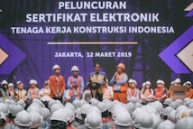 A group of people wearing construction helmets and safety vests gather on stage for an event. A speaker stands at a podium, delivering a speech in front of a large crowd. The background features a large display with text in a foreign language, set against a backdrop suggesting a construction theme.
