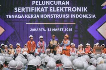 A group of people wearing construction helmets and safety vests gather on stage for an event. A speaker stands at a podium, delivering a speech in front of a large crowd. The background features a large display with text in a foreign language, set against a backdrop suggesting a construction theme.