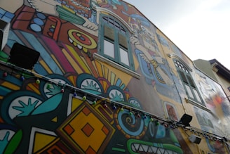 A vibrant mural on the side of a building features intricate designs and bold colors. The artwork includes geometric patterns, abstract shapes, and figures reminiscent of indigenous or ancient motifs. Multicolored string lights hang along the mural, adding an additional layer of color and decoration.
