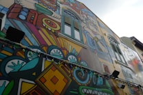 A vibrant mural on the side of a building features intricate designs and bold colors. The artwork includes geometric patterns, abstract shapes, and figures reminiscent of indigenous or ancient motifs. Multicolored string lights hang along the mural, adding an additional layer of color and decoration.
