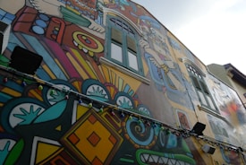 A vibrant mural on the side of a building features intricate designs and bold colors. The artwork includes geometric patterns, abstract shapes, and figures reminiscent of indigenous or ancient motifs. Multicolored string lights hang along the mural, adding an additional layer of color and decoration.