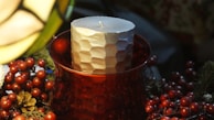 A textured cylindrical candle placed in a red glass holder surrounded by decorative elements, including pine cones and red berries. The setting is dimly lit, creating a warm and cozy ambiance.