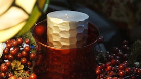 A textured cylindrical candle placed in a red glass holder surrounded by decorative elements, including pine cones and red berries. The setting is dimly lit, creating a warm and cozy ambiance.