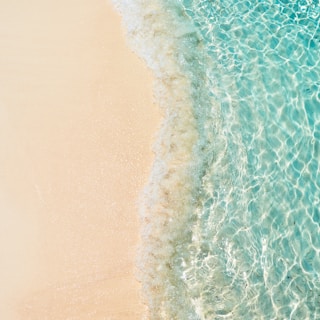 Clear turquoise water gently laps onto a sandy beach, creating a serene and tranquil coastal scene. The sunlight sparkles off the shallow waves, giving the image a fresh and inviting appearance.