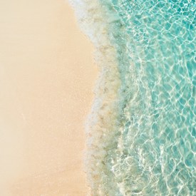 Clear turquoise water gently laps onto a sandy beach, creating a serene and tranquil coastal scene. The sunlight sparkles off the shallow waves, giving the image a fresh and inviting appearance.