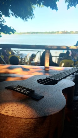 A ukulele rests on a wooden surface with a serene outdoor setting in the background. The scene includes a body of water and distant foliage, creating a peaceful and relaxed atmosphere.