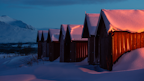 A row of small wooden cabins stands under a blanket of snow, with a warm orange light glowing from inside the cabins. Snow-capped mountains are visible in the background under a twilight sky.