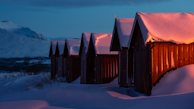 A row of small wooden cabins stands under a blanket of snow, with a warm orange light glowing from inside the cabins. Snow-capped mountains are visible in the background under a twilight sky.