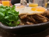 A close-up shot of a skillet containing a cooked dish with a fried egg on top. Next to the egg are tender pieces of meat, and fresh green leaves are placed on the side. In the background, there are blurred images of glasses filled with orange juice and other table items.