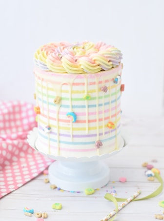 A tall, round cake decorated with pastel rainbow stripes and swirl frosting on top. The cake is adorned with small colorful cereal pieces and has a drip effect on the sides. It is placed on a white cake stand with a pink polka dot cloth in the background.