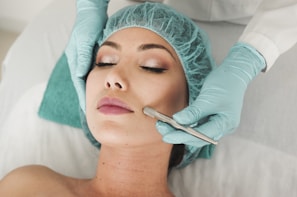 A person lies on a medical bed, wearing a surgical cap and having their skin treated with a metal instrument by a professional in gloves. The atmosphere is clinical and focused on skincare.