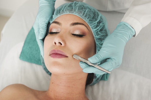 A person lies on a medical bed, wearing a surgical cap and having their skin treated with a metal instrument by a professional in gloves. The atmosphere is clinical and focused on skincare.