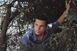 A man is surrounded by dense foliage, appearing to look contemplatively into the distance. He wears a blue, patterned shirt and has one hand gripping a tree branch, suggesting he is in a wooded or natural environment. The lighting creates a contrast between shadows and highlights, adding depth to the scene.