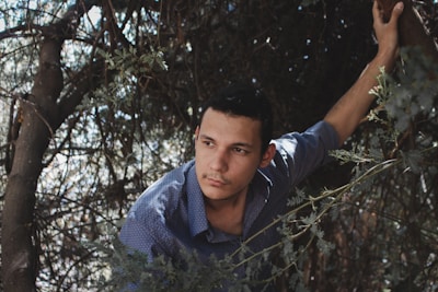 A man is surrounded by dense foliage, appearing to look contemplatively into the distance. He wears a blue, patterned shirt and has one hand gripping a tree branch, suggesting he is in a wooded or natural environment. The lighting creates a contrast between shadows and highlights, adding depth to the scene.