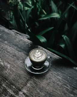 A glass cup of matcha latte sits on a wooden surface, surrounded by lush green foliage. The latte features intricate latte art on its surface, showcasing a leaf pattern.