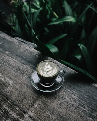 A glass cup of matcha latte sits on a wooden surface, surrounded by lush green foliage. The latte features intricate latte art on its surface, showcasing a leaf pattern.