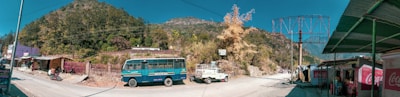 A scenic roadside view featuring a mountainous landscape with dense greenery. A blue bus and a vehicle towing a cart are parked along the road. Small shops and buildings line the street, with one displaying a large Coca-Cola advertisement. The road curves, heading into the distance toward the mountains.