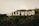 Large white letters spelling out 'HOLLYWOOD' are prominently displayed on a lush green hillside. The iconic sign stands amidst a natural landscape with scattered trees and shrubbery. The sky appears overcast, casting soft lighting over the scene.