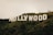 Large white letters spelling out 'HOLLYWOOD' are prominently displayed on a lush green hillside. The iconic sign stands amidst a natural landscape with scattered trees and shrubbery. The sky appears overcast, casting soft lighting over the scene.