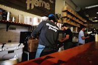 Several people are working behind a food or beverage counter in what appears to be a cafe or restaurant. They are surrounded by shelves stocked with containers and bags. One person is wearing a sweatshirt with 'Homestate Family' printed on the back, and there are various kitchen equipment and supplies visible.