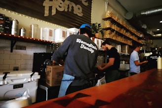 Several people are working behind a food or beverage counter in what appears to be a cafe or restaurant. They are surrounded by shelves stocked with containers and bags. One person is wearing a sweatshirt with 'Homestate Family' printed on the back, and there are various kitchen equipment and supplies visible.