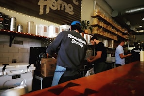 Several people are working behind a food or beverage counter in what appears to be a cafe or restaurant. They are surrounded by shelves stocked with containers and bags. One person is wearing a sweatshirt with 'Homestate Family' printed on the back, and there are various kitchen equipment and supplies visible.