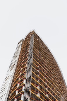 A tall, modern building under construction with numerous floors. Scaffolding is wrapped around the entire exterior, creating a grid-like pattern. The structure stretches upwards with visible structural beams and platforms.