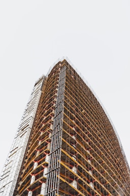 A tall, modern building under construction with numerous floors. Scaffolding is wrapped around the entire exterior, creating a grid-like pattern. The structure stretches upwards with visible structural beams and platforms.