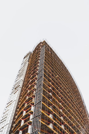 A tall, modern building under construction with numerous floors. Scaffolding is wrapped around the entire exterior, creating a grid-like pattern. The structure stretches upwards with visible structural beams and platforms.