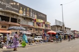 A bustling street market scene with people walking and browsing various stalls. The market is situated in front of a row of buildings, including one with a large billboard advertising perfumes. Vendors are displaying a range of goods under umbrellas and makeshift awnings.