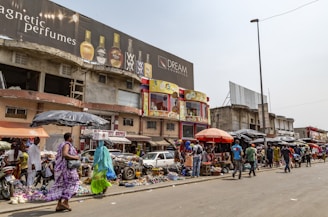 A bustling street market scene with people walking and browsing various stalls. The market is situated in front of a row of buildings, including one with a large billboard advertising perfumes. Vendors are displaying a range of goods under umbrellas and makeshift awnings.