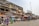 A bustling street market scene with people walking and browsing various stalls. The market is situated in front of a row of buildings, including one with a large billboard advertising perfumes. Vendors are displaying a range of goods under umbrellas and makeshift awnings.