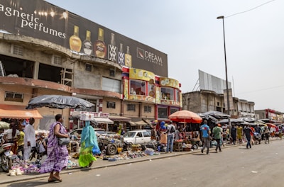 A bustling street market scene with people walking and browsing various stalls. The market is situated in front of a row of buildings, including one with a large billboard advertising perfumes. Vendors are displaying a range of goods under umbrellas and makeshift awnings.