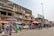 A bustling street market scene with people walking and browsing various stalls. The market is situated in front of a row of buildings, including one with a large billboard advertising perfumes. Vendors are displaying a range of goods under umbrellas and makeshift awnings.