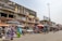 A bustling street market scene with people walking and browsing various stalls. The market is situated in front of a row of buildings, including one with a large billboard advertising perfumes. Vendors are displaying a range of goods under umbrellas and makeshift awnings.