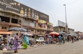 A bustling street market scene with people walking and browsing various stalls. The market is situated in front of a row of buildings, including one with a large billboard advertising perfumes. Vendors are displaying a range of goods under umbrellas and makeshift awnings.