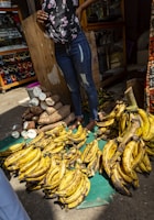 A street market scene with a person standing behind a display of ripe yellow plantains and cut cassava roots. The person is wearing jeans and a floral top. Jewelry is visible in a glass display case in the background.