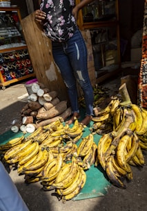 A street market scene with a person standing behind a display of ripe yellow plantains and cut cassava roots. The person is wearing jeans and a floral top. Jewelry is visible in a glass display case in the background.