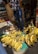 A street market scene with a person standing behind a display of ripe yellow plantains and cut cassava roots. The person is wearing jeans and a floral top. Jewelry is visible in a glass display case in the background.