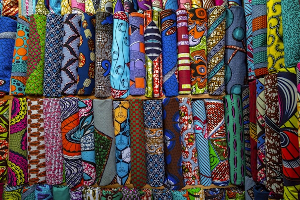A vibrant collection of various African wax print fabrics, neatly arranged in rows. Each piece features a unique pattern, including geometric shapes, swirls, and florals, in a wide array of colors like blue, orange, yellow, and green.