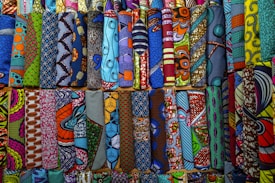A vibrant collection of various African wax print fabrics, neatly arranged in rows. Each piece features a unique pattern, including geometric shapes, swirls, and florals, in a wide array of colors like blue, orange, yellow, and green.