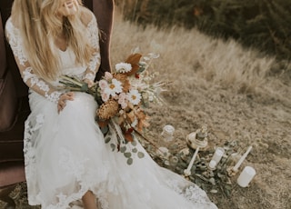 woman in white bridal gown sitting on brown wing chair while holding bouquet