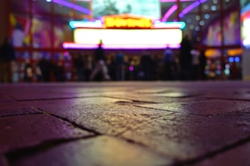 The image features a low-angle shot of a paved surface leading up to a brightly lit building, possibly a theater or cinema with vibrant neon lights. The lights create a colorful, blurred background with silhouettes of people standing near the entrance.