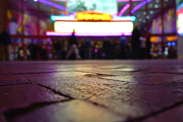The image features a low-angle shot of a paved surface leading up to a brightly lit building, possibly a theater or cinema with vibrant neon lights. The lights create a colorful, blurred background with silhouettes of people standing near the entrance.