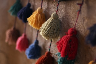 Colorful tassels made of yarn hang closely together against a textured wall. The tassels vary in colors, including red, green, blue, yellow, and orange, and appear to be handcrafted.